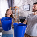 Couple Using Bucket For Collecting Water Leakage From Ceiling And Calling Plumber On Cellphone